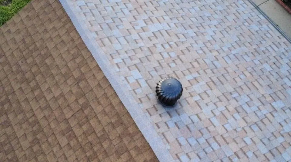 Two adjoining asphalt shingle planes in tan and light gray meeting at a sloped transition with a black roof vent