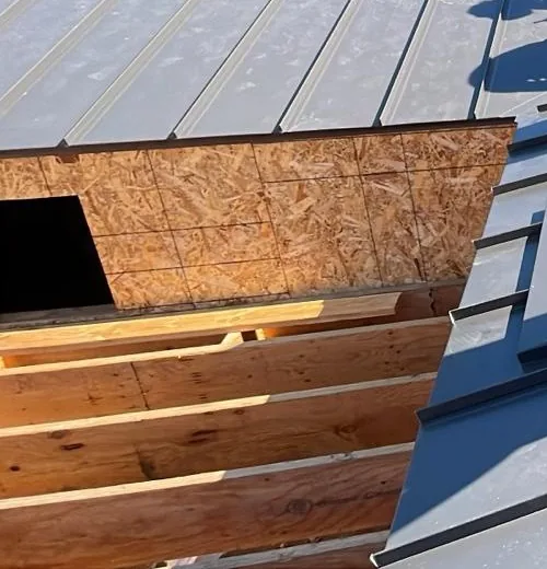Dark gray standing-seam metal roof panels beside OSB roof decking with rectangular openings.