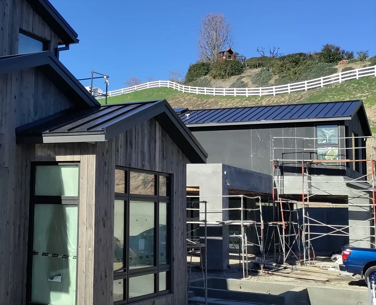 Dark standing-seam metal roofs on dark wood-sided buildings, with scaffolding, a white fence and a hillside behind.